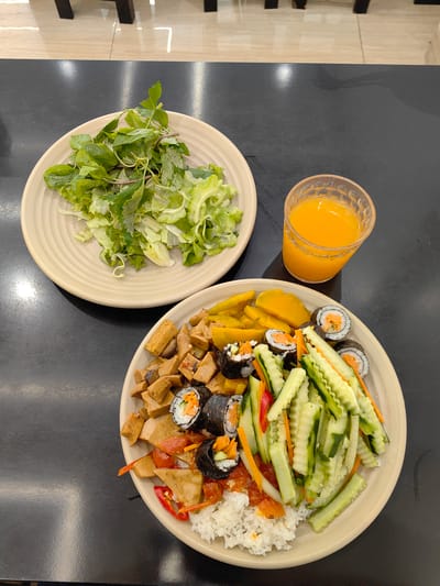 A high-angle shot shows two plates of food and a glass of orange juice on a black surface, possibly a table, in what appears to be a restaurant setting. The location is implied to be Hong Gai Ward, Vietnam.
The plate on the left contains a mix of green leafy vegetables, seemingly a salad. The plate on the right holds a more varied dish. There's white rice, pieces of what appears to be fried tofu, sliced cucumber, sliced red pepper, slices of mango and several sushi rolls. A clear plastic glass next to the plate on the right contains orange juice. The table surface is dark and appears to be smooth. The background is somewhat blurred but seems to show a tile floor with dark-colored table legs. No people are visible in the shot, and the scene depicts a static setting of food laid out.