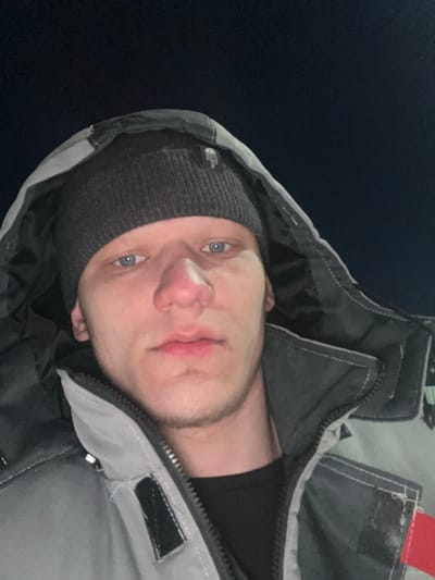 A close-up, upward-angled selfie depicts a young male individual located in the city of Залесная, Russia. The subject has light blue eyes, a pronounced nose, and light stubble visible on the chin and jawline. He is wearing a dark grey knit beanie with a small logo on the front and a grey and dark grey hooded jacket. The jacket features a zipper closure, lighter grey sections on the upper torso and arms, and a red/orange strip visible on the lower right sleeve. A black t-shirt is visible beneath the jacket. The individual maintains a neutral expression, looking directly at the camera. The background is uniformly dark, indicating a nighttime setting with no discernible environmental features.