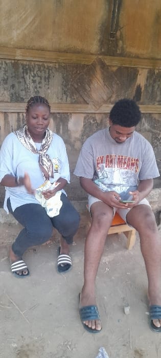 In Ashipa, Nigeria, a daylight scene depicts two individuals seated against a weathered, discolored light-brown wall with a horizontal ledge and a dark, diamond-shaped decorative element. On the left, a female with braided hair wears a light blue t-shirt, dark pants, and black-and-white striped slide sandals. A patterned scarf is draped around her neck. She holds light-colored fabric or paper in her left hand and gestures with her right hand, looking downwards. On the right, a male with dark, curly hair sits on a small wooden stool with an orange top. He wears a grey graphic t-shirt (partially reading "BY ANY MEANS"), light shorts, and dark slide sandals. He is focused on a green-cased smartphone held in both hands, looking downwards. The ground beneath them is light brown dirt or sand.
