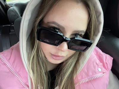 An overhead, close-up shot captures a young adult, likely female, positioned upside down from the viewer's perspective, possibly taking a selfie. Her long, straight blonde and light brown hair cascades around her face. She is dressed in a vibrant pink puffer jacket, featuring a white zipper and a silver snap button on the shoulder, paired with a light beige or off-white hoodie.
Her face is largely obscured by stylish black, rectangular sunglasses, though one light-colored eye with prominent dark eyelashes is visible above the left lens. Her eyebrows are well-defined. A small, silver hoop ring pierces her right nostril. Her lips are slightly parted, showcasing a natural pink shade. The expression appears neutral.
The setting is an interior space, specifically a car, evidenced by the dark upholstery visible to the left and elements of a car seat or seatbelt system on the right. No external environment is visible. The image conveys no specific time of day or weather. No text is visible within the frame. This scene takes place in Chituk, Russia.