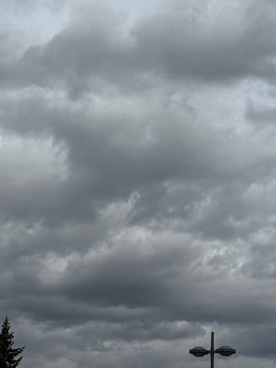 This image depicts a scene in Oberhausbergen, France, under a heavily overcast sky dominated by dense, grey stratocumulus clouds. The lower left corner shows the dark green, pointed top portion of a conifer tree. In the lower right quadrant, a vertical grey street lamp post is visible, featuring a horizontal crossbar supporting two circular, dish-like light fixtures. No people or discernible activities are present within the frame.