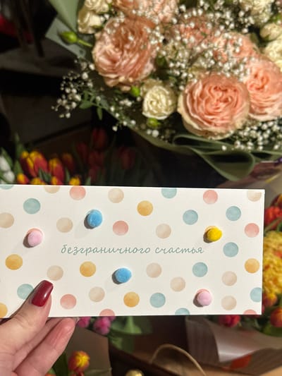 A close-up image taken in Базарово, Russia, depicts a left hand with dark red manicured fingernails holding a white greeting card in the lower-left quadrant. The card features a pattern of watercolor-style polka dots in various pastel shades (pink, blue, yellow, beige) and is adorned with small, fuzzy pom-poms in light pink, light blue, and yellow. The Russian text "безграничного счастья" (unlimited happiness) is printed in teal script on the card. In the blurred background, a large bouquet of light peach or pink roses mixed with white baby's breath, wrapped in a dark green paper or fabric, occupies the upper portion of the frame. Additional out-of-focus floral arrangements, including red and yellow tulips, are visible in the background, suggesting a flower shop or similar retail environment.