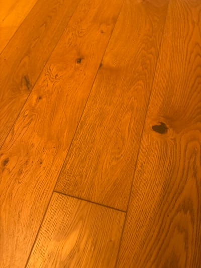 The image shows a close-up, oblique view of a wooden floor with a warm, polished finish. The planks are oriented diagonally from the bottom left to the top right. The wood grain is clearly visible, with variations in color and texture. There are several knots and imperfections in the wood, adding to its character. The lighting creates a warm glow, with highlights and shadows accentuating the surface. There are no people or other objects visible in the image.
This image is a general view of a wooden floor and does not contain any specific information that would allow for its location to be identified in the city of Herzliya, Israel.