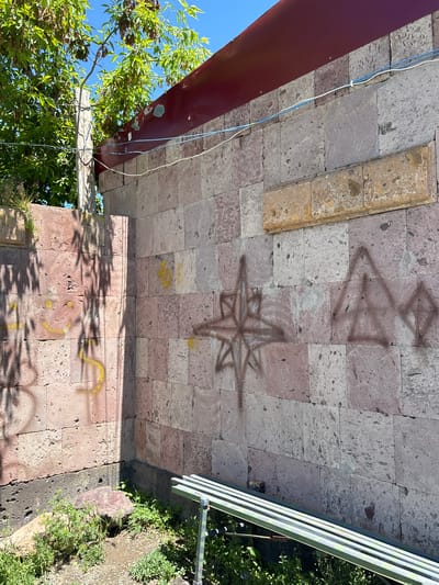 An exterior, ground-level, medium shot captures a corner of a stone building in Jrarat, Armenia, on a sunny day. The building's walls are constructed from large, rectangular, pinkish-grey volcanic stones. The lower portion of the corner wall on the left exhibits some green weeds and a reddish-brown boulder. The main wall, to the right, is adorned with spray-painted graffiti. A dark brown spray-painted eight-pointed star is visible in the center, and to its right, a triangular shape with a diamond inside is also spray-painted in dark brown. Faint yellow markings are present on the left side of the wall, including a curvilinear mark and a symbol resembling a "4". Above the stone wall, a dark red metal roof overhang is visible, with wires running horizontally across the building. Trees with green leaves and brown seed pods are seen in the upper left corner, casting shadows on the wall. A green metal bench with a slatted seat is positioned in the lower right foreground, partly obscured by weeds. The ground in front of the bench is dirt with scattered green vegetation. The sky is a clear blue.