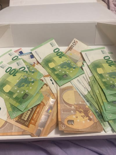 The image shows a collection of European banknotes, primarily 100 Euro notes and some 50 Euro notes, spread out inside a white box. The banknotes are arranged in a somewhat disordered manner, with some overlapping and fanned out. The lighting suggests an indoor setting, possibly a home or an office. The banknotes appear to be genuine currency. There is no indication of the location in Novi Petrivtsi, Ukraine. The image focuses on the money itself.