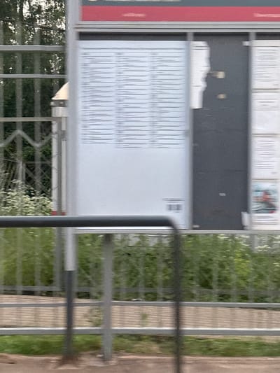 The image shows a blurred view of a timetable display at a train station in Povarovo, Russia. The timetable is mounted on a white board with some parts obscured by a dark panel. There is a red header at the top of the display. The foreground is partially obscured by a metal railing. Beyond the railing, there is lush green vegetation. The overall impression is of an outdoor setting, likely a train station platform, with the timetable serving as the central subject.