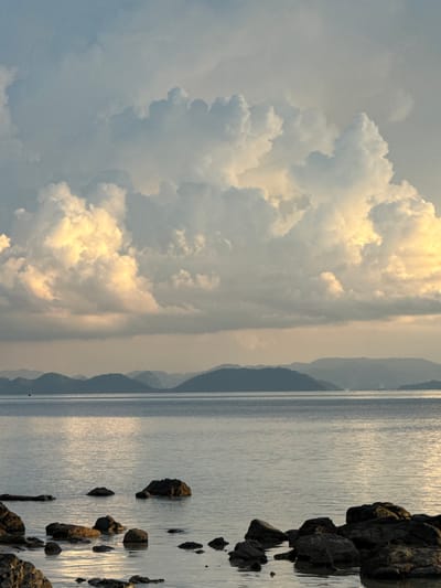 A vertical, wide shot captures a tranquil seascape with dramatic clouds at sunset. The foreground is dominated by dark, wet rocks partially submerged in calm, reflective water. The water stretches to the horizon, where a series of layered, hazy mountains recede into the distance. Above the mountains, the sky is filled with large, voluminous cumulus clouds, illuminated from behind by the setting sun, creating a warm, golden glow on their undersides, contrasting with the cooler, blue-grey tones of the upper clouds. The overall atmosphere is serene and majestic. The image is taken from the coast of Baan Pang Ka, Thailand, a location known for its picturesque bays and islands.