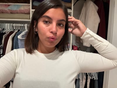 An adult female is depicted in a close-up selfie, posing indoors within what appears to be a closet or dressing area. She has dark, shoulder-length hair, a tanned complexion, and is wearing a light-colored, long-sleeved ribbed top. Her lips are puckered, and her gaze is directed at the camera. Two silver hoop earrings are visible on her left ear. Her right hand is positioned near the right side of her head. The background consists of an organized closet interior featuring multiple white shelves and hanging rods. The left side displays folded textiles in various colors, including pink and blue, on upper shelves, and numerous garments on white hangers below. The right side reveals a white, fluffy coat hanging, a dark red garment, and small pink and brown bags resting on a lower shelf. A white door frame is partially visible on the far right. The specific location in the city of Princeton, United States, cannot be determined from the provided image.