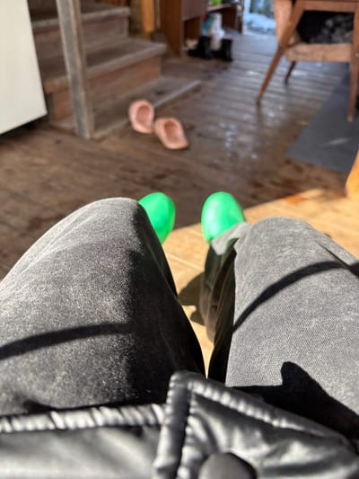 The image shows a downward-angled view of a person's legs, clad in gray jeans, with bright green clogs on their feet. Part of a black jacket is visible on the right side of the frame. The person appears to be standing on a wooden surface, possibly a porch or deck. In the background, there are wooden stairs leading down, with a pair of light pink clogs resting on one of the steps. The image's location is implied to be in Zheleznodorozhnyi, Russia, based on the prompt's instructions, but this is not verifiable from the image itself.