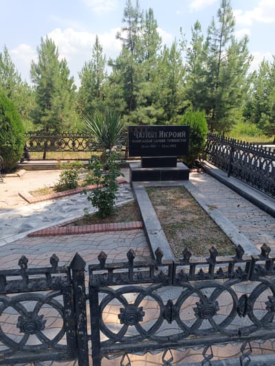 The image shows a gravestone in a cemetery. The gravestone is black with writing on it in what appears to be Cyrillic script. Behind the gravestone are several evergreen trees. A black metal fence surrounds the grave. The ground in front of the grave is paved with stone. A small rose bush is planted to the left of the grave. The overall scene is calm and well-maintained. The gravestone is inscribed with "Чалол Икромий" and dates "20.09.1909 - 10.04.1993". This is the burial site of Jalol Ikromi, a prominent Tajik writer, located in Dushanbe, Tajikistan.
