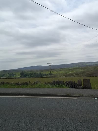 An overcast, cloudy sky looms over a rolling green landscape in Barnsley, United Kingdom. In the foreground, a section of dark asphalt road is visible, bordered by a low, rough stone wall. Beyond the wall, a field of grass and some vegetation stretches out. Further back, a line of trees and a telephone pole with wires can be seen, leading towards a series of undulating hills that recede into the distance. The overall impression is of a rural, somewhat muted scene on a cloudy day.
