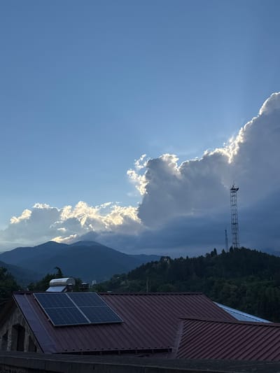 This is a vertical, medium-shot photograph taken outdoors. The scene depicts a mountainous landscape with a clear blue sky dominated by dramatic clouds. A cell tower is visible against the cloudy sky, and a forest covers the mountainside in the background. In the foreground, the roof of a building is visible, featuring two solar panels and a water heater.

The time of day appears to be late afternoon or early evening, as suggested by the angled sunlight illuminating the edges of the clouds and casting a darker hue over the mountains. The weather is partly cloudy, with large, textured clouds creating a dynamic contrast with the clear sky.

The location cues, such as the mountainous terrain and the style of architecture (red corrugated roof), are consistent with a rural or semi-rural setting in a mountainous region like Dilijan, Armenia. There are no people or specific activities depicted, creating a sense of serene, natural beauty. The image focuses on the interplay of natural elements and human infrastructure against a scenic backdrop. No text is visible in the image.