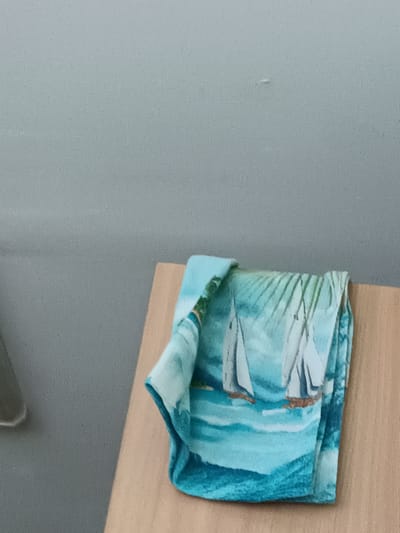 A close-up, eye-level shot shows a folded blue and white towel with a sailboat pattern draped over a light-brown wooden surface. The towel is positioned on the right side of the frame, with its pattern of sailboats on a blue ocean filling the lower half of the image. The background is a textured, light gray wall. The scene is indoors, likely in a residential setting, and there are no people or animals present. The lighting is soft and diffused. The overall impression is one of calm and domestic simplicity. The location in the city of Ключи, Russia is not discernible from the image.