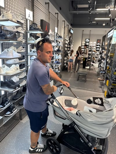 In Carini, Italy, a man stands in a shoe store, pushing a baby stroller. He is wearing a purple t-shirt, dark blue shorts, and black Adidas sneakers with white stripes. He has tattoos on his arms and legs. The stroller is gray and has a white baby inside. The store is brightly lit and filled with shelves of sneakers. In the background, other shoppers browse the merchandise. The store appears to be a large, modern retail space with a clean, industrial aesthetic.