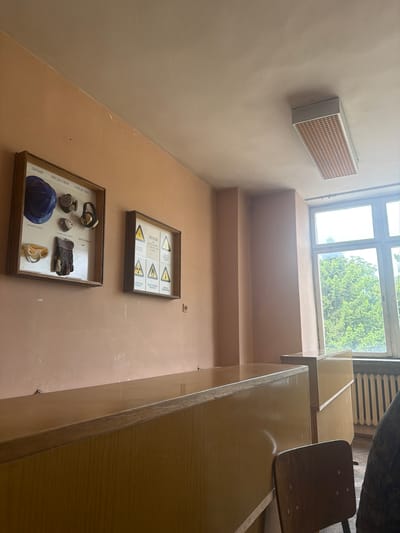 The image depicts an interior space, likely a building in Dolna Mitropolia, Bulgaria. The walls are peach-colored, and there is a window with a view of green trees outside. On the wall are two framed displays: one appears to contain safety equipment, including a blue helmet, gloves, and respirators, while the other contains radiation hazard symbols. A rectangular ventilation unit is mounted high on the wall. Desks and a chair occupy the right side of the frame. The perspective is from a low angle, looking upwards towards the window.