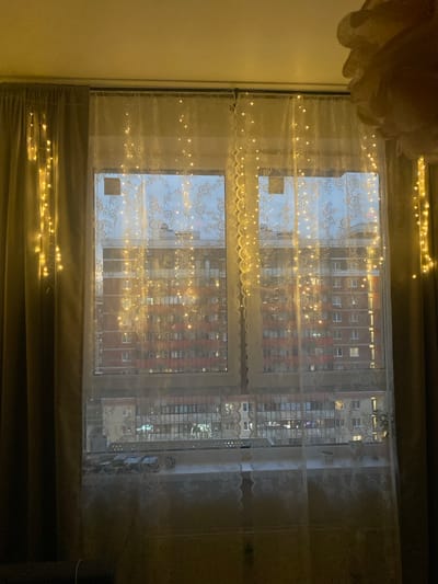 A medium shot captures a window adorned with string lights, overlooking a city apartment building at dusk. The window is covered by two layers of curtains: heavy, dark curtains on the sides and a sheer lace curtain in the center. String lights with small, yellow bulbs are draped along the edges of the window and across the lace curtain, casting a warm glow. The apartment building visible through the window has a brick facade and multiple stories, with numerous windows showing interior lights. A faint hint of a street or the ground is visible at the bottom of the frame, suggesting an urban setting. The overall scene conveys a cozy, festive atmosphere.