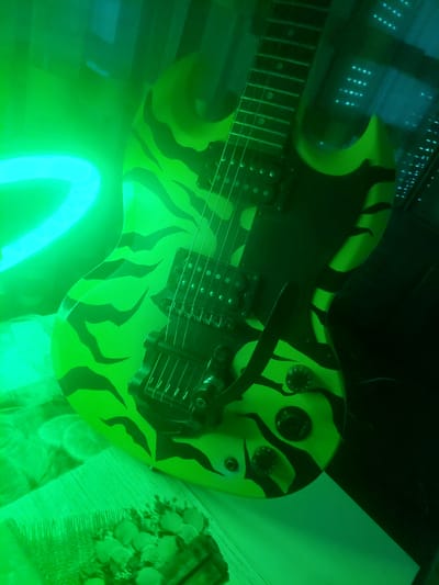 An electric guitar with a neon green and black tiger stripe pattern is displayed on a surface. The guitar has two pickups and a vibrato system. The scene is illuminated by neon green and blue lights, creating a vibrant and somewhat abstract atmosphere. The background appears to be a dimly lit room with some vertical elements that could be blinds or shelves. There are no people present in the image. The location of Markovo, Bulgaria is not discernible from the image.