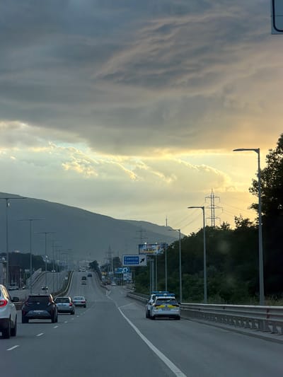 This image depicts a scenic view of a highway in Pancharevo, Bulgaria, during a dramatic sunset. The sky is filled with a mix of dark, ominous clouds and bright, golden light, suggesting a recent or impending storm. The sun is low on the horizon, casting a warm glow that illuminates the clouds and the landscape.
The highway stretches into the distance, with several cars visible. In the foreground, a police car with flashing blue lights is stopped on the right side of the road, possibly attending to an incident or directing traffic. Further down the highway, other vehicles, including a silver SUV and a dark sedan, are also visible.
On the left side of the highway, a lush green hillside is covered in trees. In the background, a large mountain range dominates the skyline, shrouded in a dusky haze. Power lines and transmission towers are scattered across the landscape, hinting at the infrastructure supporting the area.
Several road signs are present. One sign with blue lettering indicates "Самоков Samokov," a town located in Bulgaria, providing a geographical cue. Another sign features a speed limit of 120 km/h. A billboard advertising "THE BEST" is also visible on the left.
The overall impression is one of a dynamic and ever-changing natural environment juxtaposed with the orderly flow of traffic on a modern highway. The weather conditions, with the dramatic interplay of light and shadow, create a sense of awe and perhaps a touch of foreboding. The presence of the police car suggests that some event, perhaps an accident or a traffic stop, is taking place. The image captures a moment of transition, both in terms of the time of day and potentially the unfolding events on the road.