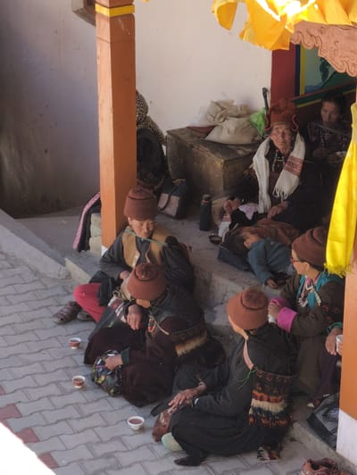 The image depicts a group of people, likely women, gathered outdoors on a paved walkway in Pibiting, India. The individuals are seated on steps and the ground, some appearing to be in conversation. Several people are wearing traditional clothing and headwear, including distinctive red hats. Three small cups containing a dark liquid are placed on the ground in front of some of the seated individuals. The environment suggests a sunny day, with shadows indicating overhead structures. A portion of an orange pillar is visible on the right side of the frame, and yellow fabric hangs above the scene. The background includes a white wall and what appears to be a wooden chest. The overall atmosphere is informal and communal.
