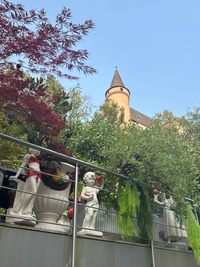 The image captures a view from an elevated position, looking towards a castle tower in Vianden, Luxembourg. In the foreground, a metallic railing runs horizontally across the frame, separating the viewer from decorative sculptures and lush greenery.

On the left, a white statue of a female figure, adorned with red fabric around her waist and chest, stands beside a large, ornate white vase filled with artificial fruits. To the right of the vase is a white statue of a cherubic child holding a red object in its hands and a basket of flowers. Further to the right, another white statue of a female figure in a flowing dress is visible, holding a red apple. Cascading green ferns hang from above, adding to the vibrant foliage that surrounds the statues.

Behind the statues and railing, a dense arrangement of trees and shrubs creates a natural backdrop. Prominent among these is a tree with striking deep red, maple-like leaves, contrasting with the lighter green of other trees.

In the background, the distinctive conical tower of Vianden Castle rises above the foliage, its orange stone structure capped with a dark, pointed roof. The castle appears to be situated on a hill, with a clear, light blue sky overhead. The overall scene suggests a decorative garden or terrace area with a view of the historic castle.