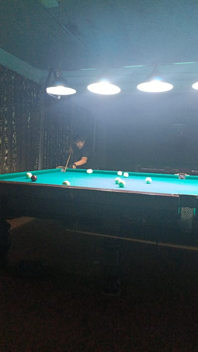 An indoor scene depicts a man playing billiards. The man, wearing a dark t-shirt and black gloves on his left hand, is positioned behind a pool table with a green felt surface. He is holding a cue stick with his right hand, aiming at a white ball. Several white billiard balls are scattered on the table, with one red billiard ball also present. The pool table is illuminated by overhead lights. The background features dark curtains and a dark wall. The overall lighting suggests a dimly lit environment.
