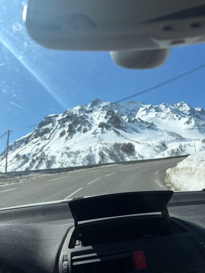 The image, taken from inside a vehicle, shows a view of the French Alps, specifically near Le Monêtier-les-Bains, France. A snow-covered mountain dominates the scene through the car's windshield. Outside of the vehicle is a road. Inside the vehicle is a dashboard and rearview mirror. The sky is blue. Overhead wires are visible.