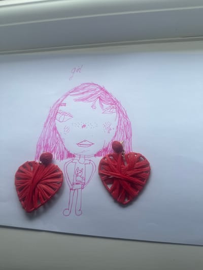 The image shows a pink pen drawing of a girl on a white sheet of paper. The drawing depicts the girl with pink hair, freckles, and a shirt with a cat design. To the left and right of the drawing are two red heart-shaped earrings. The earrings are made of woven red material and have a gold post. The paper is lying on a light-colored surface, possibly a desk or table, with a white element at the top of the frame, which could be a windowsill or shelf. The image is taken from a top-down perspective. There is no information in the image to suggest a location in Wellingborough, United Kingdom.