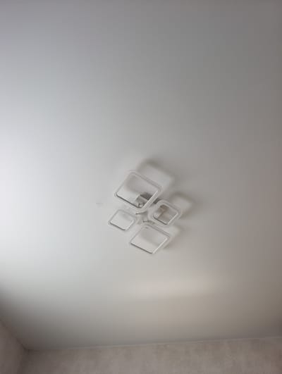 The image shows a modern ceiling light fixture with four interconnected square frames mounted on a white ceiling. The fixture is positioned in the upper right quadrant of the frame. The ceiling appears to be smooth and free of imperfections, with subtle shadows cast by the light fixture. The lower portion of the image shows a textured wall with a light beige or gray color. There are no people or any other objects visible in the scene. The image is a static shot of an interior space. The provided location information is not visually verifiable from the image content.