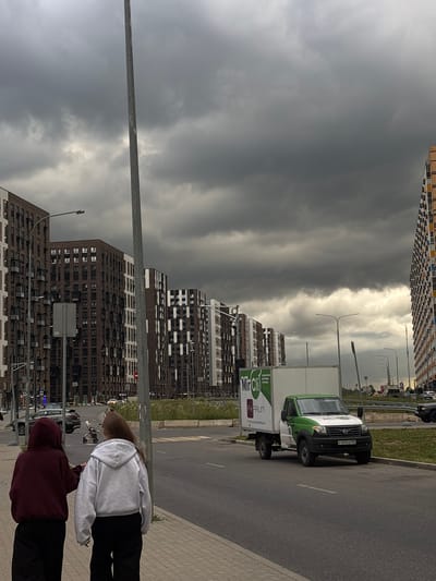 This image captures a street scene in Misaylovo, Russia. The sky is heavily overcast with dark, dramatic clouds, suggesting impending rain or a storm. In the background, tall, modern apartment buildings with a mix of dark brick and white facades dominate the skyline. A white and green delivery truck with the logo "Mr. Gilli" and the text "FEELGOOD" is parked on the asphalt road. The truck has a license plate that appears to read "P147CO 62".
In the foreground, two individuals are visible from the back, standing on a paved sidewalk. One is wearing a light grey hoodie and dark pants, while the other is in a dark red hoodie and dark pants. Their posture and the casual attire suggest a relaxed moment. The scene is likely set during the daytime, given the natural light, but the stormy weather creates a somber atmosphere. The presence of modern architecture and a delivery truck indicates an urban or suburban environment.