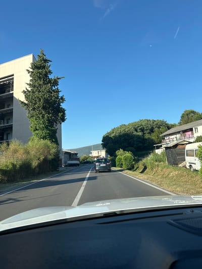 The image depicts a view from inside a vehicle traveling on a road in Bijela, Montenegro. The road is paved and marked with a white line. On the left side of the road, there is a large, light-colored building, possibly under construction, with balconies visible. A tall, mature evergreen tree stands beside the building, its branches reaching towards the clear blue sky. Dense green foliage and bushes are present along the roadside. On the right side of the road, there are houses, a smaller wooden structure, and a white van. A dark-colored car is driving ahead of the vehicle from which the photo is taken, and another car is parked further down the road on the left. The scene is illuminated by sunlight, casting shadows on the road. In the distance, hills are visible under a clear, cloudless sky.