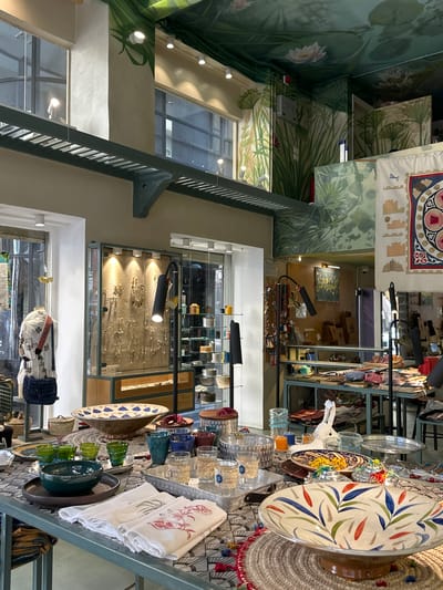 The image depicts the interior of a shop in Cairo, Egypt. The shop is decorated with murals of lush vegetation and lotus flowers. Merchandise is displayed on metal shelving. Items for sale include decorative plates, glassware, textiles, and jewelry. There are no people visible in the image. Display cases are illuminated with spotlights.