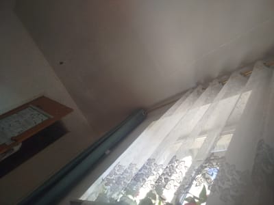 This is a photograph taken indoors, looking upwards. The ceiling occupies a large portion of the frame, with a visible seam running across it. To the left, a wooden frame with what appears to be a bulletin board or a framed document is mounted on a wall. Adjacent to this, a dark, rectangular opening suggests a doorway or a recess. A green curtain hangs down on the left side of the frame. On the right, a sheer white curtain with a floral pattern is drawn across a window, revealing the outlines of window panes and some reflections. Sunlight streams through the curtain, creating bright areas and casting shadows. The overall lighting is dim, with the exception of the areas illuminated by the sunlight. No people or specific actions are discernible. The image does not contain any elements that can identify its location within the city of Naryn City, Kyrgyzstan.