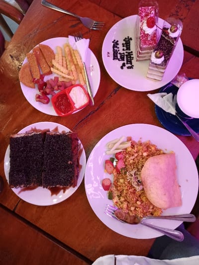 An overhead shot captures a wooden table laden with various dishes, suggesting a celebratory meal in Situbondo, Indonesia. The warm lighting casts a slightly purple hue over the scene.

In the upper left, a white plate holds a serving of golden French fries, several fried breaded fish cakes or nuggets, and cut-up sausages shaped like octopuses or flowers. Accompanying these are small red and white bowls containing ketchup and mayonnaise.

To the upper right, another white plate features two slices of cake: one appears to be a red velvet or strawberry-flavored layer cake with a cherry on top, and the other a chocolate or caramel layered cake, also topped with a cherry-like garnish. "Happy Mother's Day" is elegantly written in chocolate syrup on the plate beside the cakes. A white cup with a creamy white beverage sits on a blue saucer next to this plate.

At the bottom left, a plate presents two substantial slices of a dark, crumbly cake or bread, possibly an Indonesian specialty, generously drizzled with chocolate sauce. Its textured surface resembles shredded chocolate or a dense crumb topping.

Finally, at the bottom right, a plate is filled with a portion of fried rice (*nasi goreng*), mixed with corn, chopped green vegetables, and sliced sausages. A large, pinkish-orange *kerupuk* (cracker) leans against the fried rice, and a spoon and fork are placed beside the dish.

The overall scene conveys a casual yet festive dining experience, with a mix of savory and sweet items, including recognizable Indonesian culinary elements. The table is dark brown, showcasing a natural wood grain.