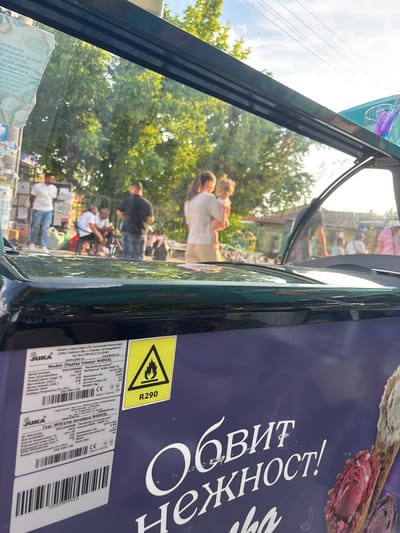 The image depicts an outdoor scene in Kostievo, Bulgaria, viewed through the transparent top panel of a dark-colored JUKA M400SL commercial display freezer. The freezer's label identifies its Ukrainian manufacturer as "Juka-invest" LTD and specifies R290 refrigerant with a corresponding flame warning symbol. Bulgarian branding text "Обвит нежност!" (Wrapped in tenderness!) is visible on the freezer's side, alongside images of ice cream cones.
The scene beyond the freezer's glass, appearing somewhat refracted, shows a group of approximately seven individuals in what appears to be a public or market area during late afternoon or early evening, indicated by warm sunlight. From left to right: a man in a white t-shirt and blue jeans stands beside stacked containers; two men are seated on the ground; a woman in light clothing holds a young child; and a man in a dark t-shirt stands facing away. Another partial figure is visible on the far right. Various bags and items are scattered on the ground near the seated individuals. The background features lush green trees, partially obscured buildings with tiled roofs, and a clear blue sky with overhead power lines. A utility pole adorned with numerous Cyrillic-script posters is visible on the far left.