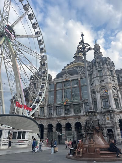 The image depicts a scene in Antwerp, Belgium, with the Antwerp Central Station prominently featured. A large Ferris wheel, labeled "The View," stands in front of the station. Several people are present in the scene. Some are seated on or near a bronze sculpture of a man, while others are walking nearby or inside the ticket booth of the Ferris wheel. The architecture of the train station is ornate and constructed of stone. A rainbow flag is visible on the side of the train station. The weather appears to be overcast.