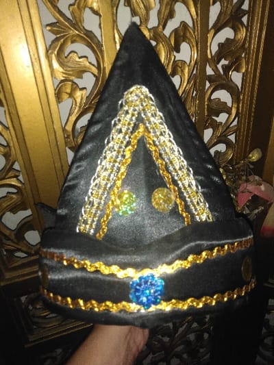 A human hand with light skin tone holds a black, conical fabric cap upwards. The cap features intricate decoration. Its upper front displays two inverted 'V' or 'A' shaped patterns composed of interwoven gold and silver braided cords. Below these, two circular, multifaceted, reflective embellishments are visible; one is greenish-yellow, the other yellow. The cap's lower band is adorned with two horizontal, wavy gold trim lines, between which a prominent, multifaceted, reflective blue gem is centrally placed. Two small, dull, circular gold-colored objects are positioned on the far right of this lower band. The background consists of an ornate, three-dimensional golden structure, exhibiting elaborate scrollwork and floral motifs, consistent with carved wood or molded metal paneling in an interior setting.
