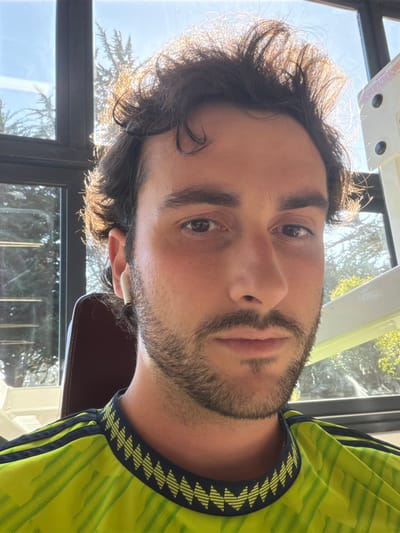 A man's close-up portrait displays his dark hair, styled with a slight wave and highlighted by sunlight. He has brown eyes and a neatly trimmed beard. He wears a yellow and green patterned shirt with dark stripes on the shoulders. Behind him are window panes, suggesting an indoor setting with an outdoor view of greenery. He is wearing a white in-ear headphone.