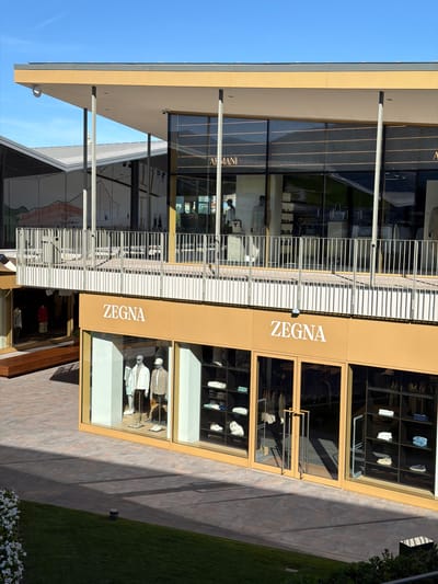 The image shows a sunlit scene of an outdoor shopping area with stores. In the foreground, a Zegna store is prominently displayed with mannequins in the window showcasing clothing. The store has large glass windows and double doors. To the left of the Zegna store, another store with mannequins is visible. Above these stores, a walkway or balcony with a metal railing overlooks the scene. In the background, another store with "ARMANI" signage can be seen through large windows. The architecture is modern, with clean lines and flat roofs. The environment is paved with stone tiles and has a grassy area with some white flowers on the left. The sky is clear and blue. This scene is located in the luxury outlet mall of Sanremo, Italy.