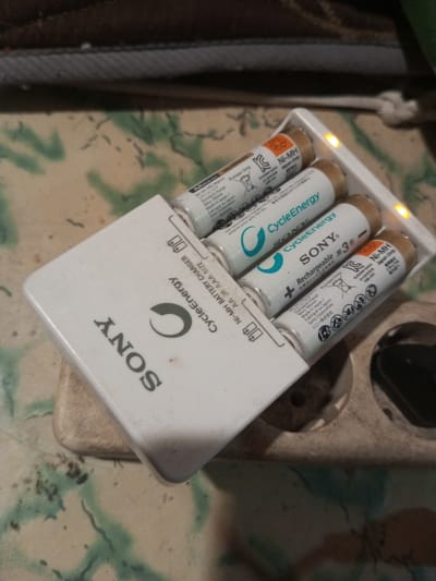 The scene, located in Bagochen Boolingpang Ueling, Bhutan, displays a white Sony CycleEnergy battery charger positioned on a beige power strip. Four cylindrical rechargeable batteries are inserted into the charger; two amber indicator lights on the charger are illuminated, indicating active charging. Two batteries bear light blue 'CycleEnergy NI-MH' branding, while the other two are white and feature 'SONY CycleEnergy Rechargeable 3' labels. The power strip exhibits multiple circular outlets, one containing a dark plug. Below the power strip and charger, a surface with a light background and an abstract green and beige pattern is visible. The upper background shows an out-of-focus, dark, textured fabric. No individuals are present.