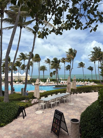 The image depicts a scene in Key Biscayne, United States of America. It features a pool area with palm trees lining the waterfront. Tables and chairs with closed umbrellas are arranged along a brick-paved path, bordered by manicured hedges. In the foreground, there's a tree with lush green leaves. The pool is flanked by lounge chairs and umbrellas, occupied by people. A standing menu board is visible on the brick path. The sky is blue with scattered clouds.