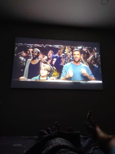 The image shows a projection of a scene from a film or show onto a screen, likely in a dimly lit room. The projected image features several people, including a young boy with light brown hair in the foreground, looking to his left. Behind him and to his right is a man with dark skin, wearing sunglasses and a tank top, holding a beverage. Further back, another man with dark hair is visible, looking intensely towards the viewer. The background of the projection is a crowd of people, some with their hands raised, creating a sense of an event or performance.
The foreground of the actual photograph reveals a dark environment, suggesting a home theater setup. Two pairs of feet are visible, one on the left and one on the right, indicating that at least two individuals are present, likely watching the projected content. The feet appear to be resting on blankets or bedding, further reinforcing the idea of a relaxed, home setting. The location in Fanipal, Belarus, is not discernible from the visual information provided in the image.