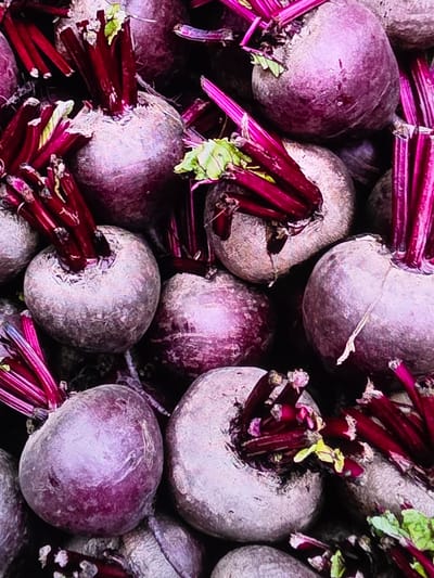 This image is a close-up, overhead shot of a large pile of fresh beets with their greens still attached. The beets are a deep purple color, and their tops are a vibrant red, some with a few small green leaves. They are tightly packed together, filling the entire frame of the image. The lighting is bright, highlighting the texture and color of the vegetables. There is no indication of the location in the city of Voluntari, Romania, nor are there any people or interactions depicted.