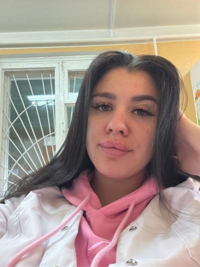 A young woman with dark hair and fair skin is pictured from the chest up, looking directly at the camera. She is wearing a pink hooded sweatshirt underneath a white lab coat, suggesting a possible medical or educational setting. Her right hand rests on her head, and her left arm is not visible. The background shows a window with metal bars and a yellow-painted wall. The lighting is bright, possibly from overhead fluorescent lights visible through the window. The image appears to be a selfie taken indoors. The location is stated as Чумна, Russia, but no specific landmarks or details in the image confirm this.
