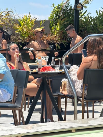 The image depicts a group of people relaxing outdoors at what appears to be a rooftop bar or restaurant in Rishon LeZion, Israel. The scene is bathed in bright sunlight, suggesting it is daytime, likely afternoon given the shadows. Lush green foliage surrounds the seating area, indicating a pleasant, possibly warm, weather condition.
Several individuals are visible, mostly partially or from the chest up. One man in the foreground wears a light-colored hat and sunglasses, appearing relaxed as he leans back. Next to him, a woman with braided hair is seen holding a piece of food to her mouth, her expression neutral. In the background, another person is holding a smartphone. The seating area features modern, dark chairs with metal accents and wooden elements, consistent with an outdoor leisure establishment. There are also drinks and some food items on a table between the people.
The overall atmosphere is casual and leisurely, with people enjoying a day out, possibly socializing or dining. The presence of a lighting fixture with a hexagonal pattern suggests that the venue might be active in the evening as well. No visible text is present in the image.