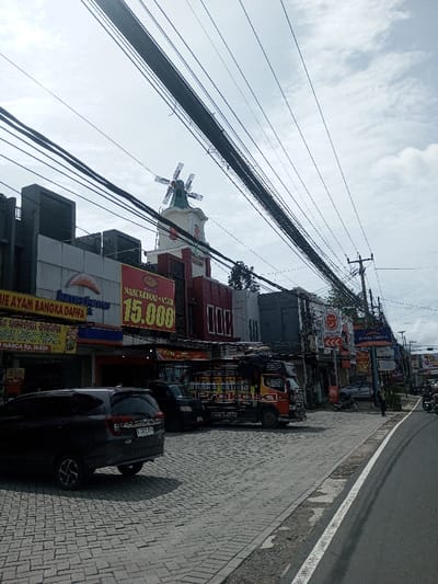 The image shows a street view with several businesses. A tall building with a windmill on top stands out. Signs advertise "Mie Ayam Bangka Dafifa" and "Nasi Kebuli + Ayam 15.000". A dark SUV is parked on a cobblestone street, and a delivery truck is visible. Overhead, a dense network of power lines crisscrosses the sky. Several storefronts, including a "Kimia Farma" pharmacy, line the street.