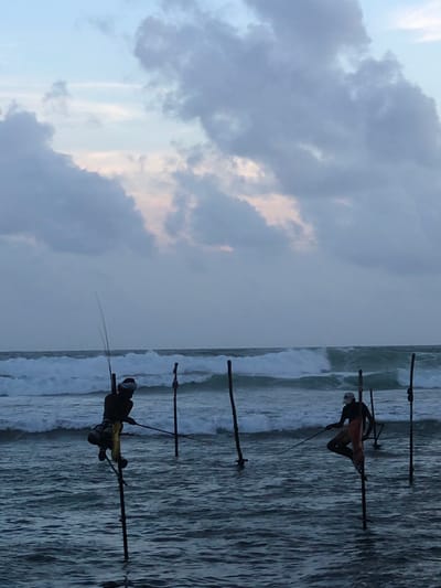 The image depicts two stilt fishermen in the shallow waters off the coast of Kathaluwa, Sri Lanka, during overcast daylight. The fishermen are perched on tall wooden stilts, each holding a fishing rod. The ocean is rough with visible waves crashing. The sky is filled with gray and white clouds, with hints of pale pink and blue. The overall scene is somber and highlights the traditional fishing practice unique to the region.