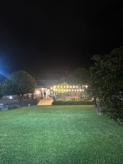 The image depicts an outdoor scene at night in Moreira, Portugal. A well-manicured lawn occupies a significant portion of the right side. A structure with multiple levels is visible, illuminated from within and by strings of decorative lights. People are present on the lighted patio of the building. Trees are interspersed throughout the scene, framing the structure and adding to the outdoor ambiance. The left side of the image is dark, contrasting with the illuminated building and lawn.
