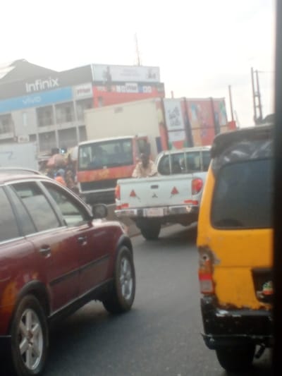 The image presents a congested urban street scene in Abule Egba, Nigeria, under diffused daylight. In the left foreground, the front-left section of a dark red SUV is visible, oriented to the viewer's left. To the right foreground, the rear-right quarter of a yellow commercial van or minibus is present, exhibiting significant rust and damage, particularly around its partially visible taillight assembly.

In the midground, a white pickup truck is centrally positioned, displaying red and white triangular reflective markers on its tailgate. An adult male, wearing a light-colored top, is visible standing or sitting within the cargo bed of this pickup truck, facing away from the viewer. Behind the white pickup, a red and white commercial truck or bus is partially obscured.

The background features multiple urban structures. On the left, a multi-story building prominently displays large "Infinix" and "vivo" mobile phone brand signage. An adjacent building has a red facade with smaller, partially legible signs, including "TOL." Further to the right, a large, colorful billboard or graphic display featuring text such as "Sprinkle Drops" is partially visible. Overhead utility lines, poles, and a distant antenna-like structure are discernible against a light, hazy sky. Vehicles appear to be stationary or in slow-moving traffic on an asphalt road. No explicit interactions between individuals are discernible.