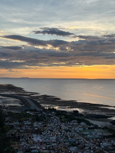 The image captures a panoramic view of a coastal landscape at sunset in the Lý Sơn Special Administrative Region, Vietnam. The sky is filled with dramatic clouds, ranging from dark gray to light orange and yellow hues, with the bright orange glow of the setting sun prominent on the horizon. The sea, calm and reflecting the sky's colors, stretches towards a distant, hazy shoreline with faint outlines of mountains.
In the foreground and mid-ground, a densely populated cemetery occupies the land adjacent to the coast. Thousands of graves, characterized by varying tombstone designs, colors (predominantly white, gray, and pastel shades), and sizes, are visible, creating a textured mosaic. The cemetery is situated on a hillside that slopes down towards the sea. A road winds through the cemetery and along the coastline, with a few vehicles and small groups of people discernible on it, indicating some activity. The coastline itself is rocky and appears to be an intertidal zone, with visible seaweed or dark mudflats. The overall scene depicts a somber yet beautiful intersection of human life, death, and the natural environment.