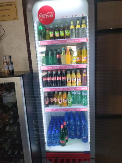 The image shows a well-stocked beverage refrigerator, likely in a bar or shop, as indicated by the "OPEN TIME" and "BAR" text in Bulgarian on a sign in the lower left. The refrigerator is brightly lit from within, showcasing a variety of bottled drinks. The primary subjects are the numerous bottles of soft drinks and water, neatly arranged on shelves. Brands like Coca-Cola, Sprite, Fanta, Schweppes, Fuze Tea, and Devin water are clearly visible. The "ICE COLD" text on the side of the refrigerator emphasizes its function.
In the background, a dark, indistinct wall and ceiling are visible, suggesting an indoor setting. The lower part of the image shows another cabinet, possibly a wine cooler, with bottles inside. The overall scene is one of commercial display and abundance of refreshments. There are no people visible in the image. The lighting suggests it could be any time of day, as it's indoors. The text "estate" is subtly visible on a dark surface below the refrigerator. The sign in Bulgarian, "ОТНО ВРЕМЕ ОБИ БАР „ПЕРМА" 8.30-22.00", translates to "OPEN TIME BAR „PERMA" 8.30-22.00", indicating the operating hours of the establishment.